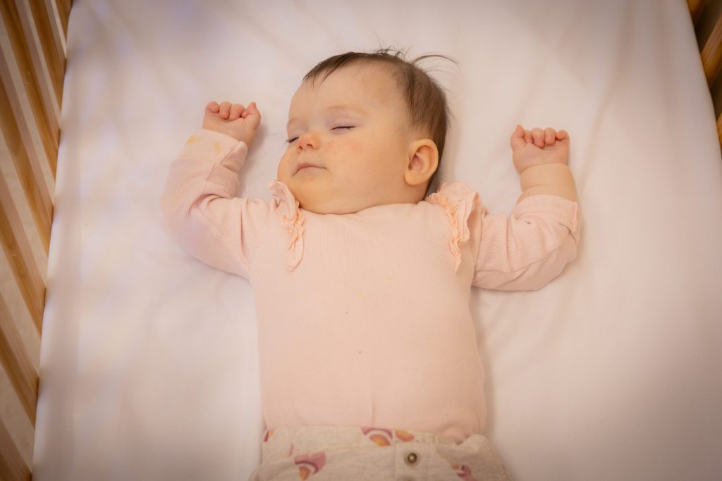 A sleeping baby lies in a crib, arms raised, wearing a light pink outfit. The setting is calm and peaceful.